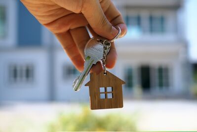 A hand holding a key with a house shaped keyring in front of a property